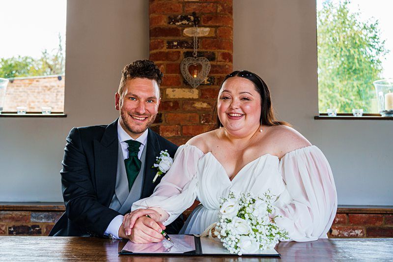 A couple in wedding attire smile at the camera while sitting at a table. The groom is wearing a dark suit with a green tie, and the bride is wearing an off-shoulder white dress and holding a white bouquet. They are posing indoors with a brick wall and windows behind them. Image by Documentary Wedding Photographer Lewis Bishop Photography.
