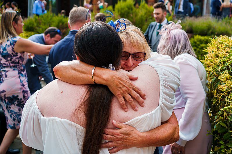 Two people embrace warmly at an outdoor gathering. The person facing the camera wears glasses and has short light hair, while the other has long dark hair and is dressed in an off-the-shoulder white outfit. Other attendees and greenery are seen in the background. Image by Documentary Wedding Photographer Lewis Bishop Photography.