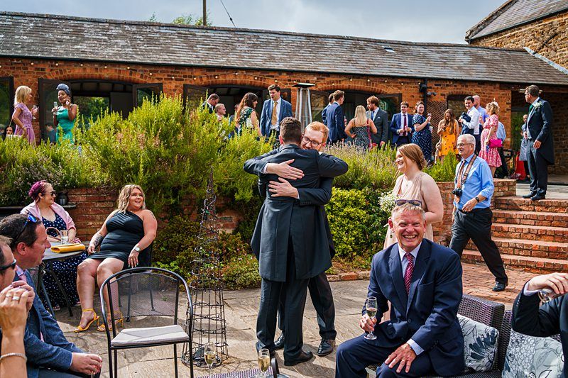 A lively outdoor wedding reception with guests mingling, laughing, and enjoying drinks. Two men in suits embrace joyfully in the center. A man in a suit sits in the foreground, smiling broadly. Stone buildings and lush greenery provide a charming backdrop. Image by Documentary Wedding Photographer Lewis Bishop Photography.