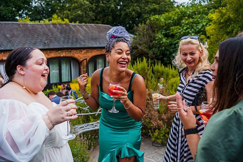 Four women are standing outside, smiling, and laughing while holding drinks. The woman in the center wears a green dress and has a red drink in her hand. They are surrounded by greenery and a brick building is visible in the background. It's a joyful moment. Image by Documentary Wedding Photographer Lewis Bishop Photography.