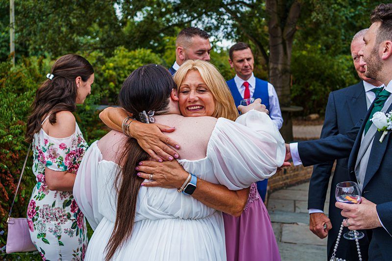 A bride in a white dress embraces a guest in a pink dress at an outdoor event. Other guests, dressed in colorful attire, chat and smile in the background. Trees and greenery create a natural setting. The atmosphere appears joyful and celebratory. Image by Documentary Wedding Photographer Lewis Bishop Photography.