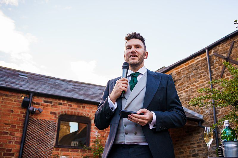 A man with curly hair is standing outdoors, holding a microphone and smiling while giving a speech. He is wearing a suit with a green tie and has a small device in his other hand. Behind him, there are rustic brick buildings and a clear sky. A bottle and glass are on a table nearby. Image by Documentary Wedding Photographer Lewis Bishop Photography.