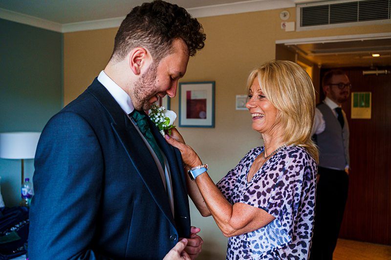 A woman pins a white flower boutonniere on a man's suit jacket while smiling. The man, dressed in a dark suit, looks down at the boutonniere. Another person stands in the background, dressed formally. The scene takes place in a warmly lit room. Image by Documentary Wedding Photographer Lewis Bishop Photography.