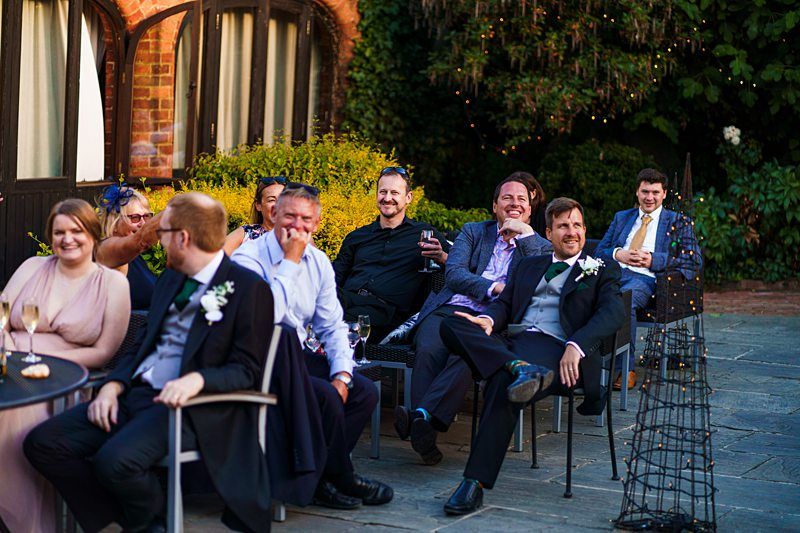 A group of well-dressed people sit and stand outdoors, enjoying what appears to be a social or formal event. Some individuals are wearing suits and ties, while others hold drinks and smile, indicating a lively and pleasant atmosphere. A brick building and greenery are in the background. Image by Documentary Wedding Photographer Lewis Bishop Photography.