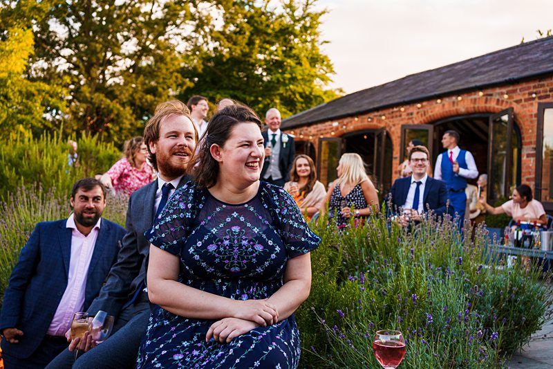 A group of people enjoying an outdoor event, seated among lavender bushes. A woman in a floral dress and a man in a suit, both smiling, are in the foreground. Others in semi-formal attire sit and stand in the background near a rustic brick building. Image by Documentary Wedding Photographer Lewis Bishop Photography.