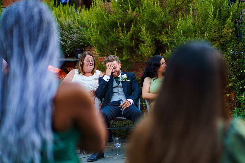 A man in a suit, seated between two women, covers his face with his hand in an emotional moment. One woman is smiling while the other looks on attentively. They are outdoors, surrounded by lush greenery, with blurred figures in the foreground. Image by Documentary Wedding Photographer Lewis Bishop Photography.