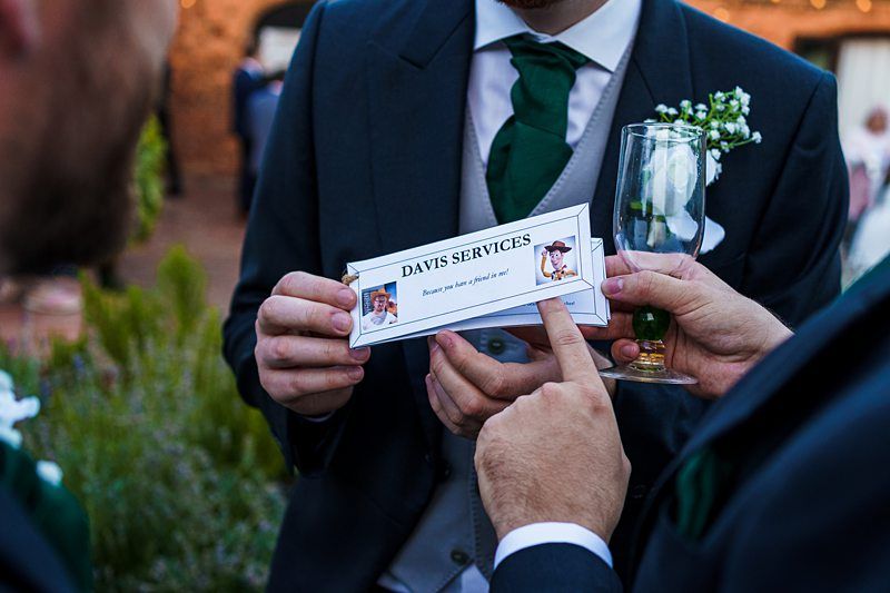 Three individuals in formal attire are holding and pointing at a business card labeled "DAVIS SERVICES" featuring images of a chef and a graduate. One person holds a champagne flute decorated with a small flower. The background shows an outdoor setting with greenery. Image by Documentary Wedding Photographer Lewis Bishop Photography.