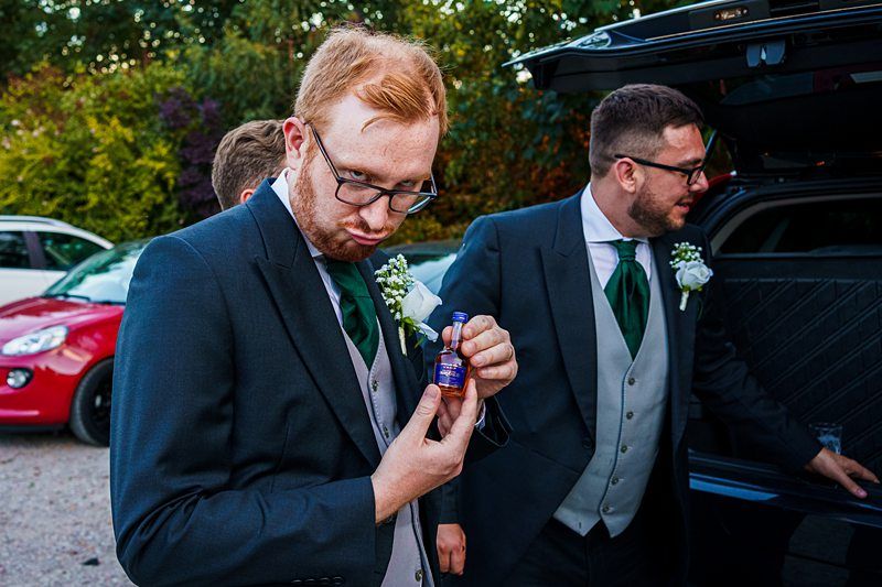Two men in suits stand near an open car trunk. The man in the foreground, wearing glasses, a dark suit, and a green tie, is making a playful expression while holding a small bottle. The man in the background, also in similar attire, is looking away. Image by Documentary Wedding Photographer Lewis Bishop Photography.