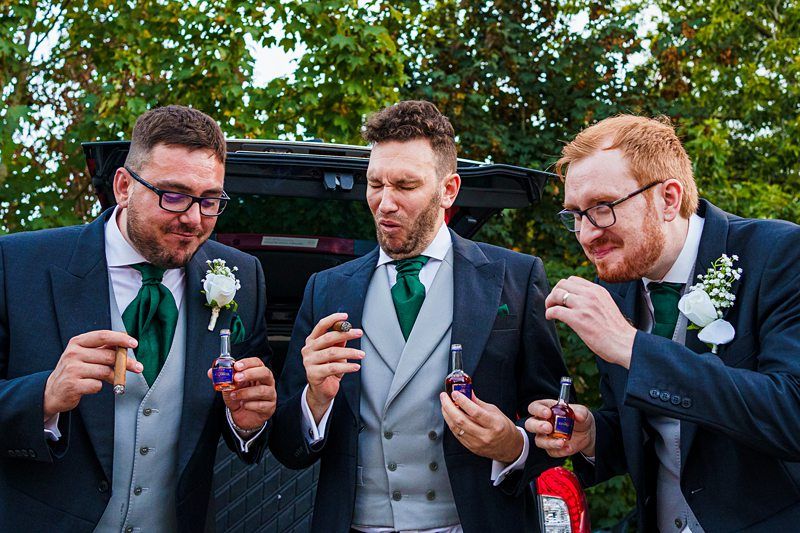 Three men in suits and green ties are standing in front of a car, each holding a cigar and a small bottle. They appear to be at a formal event, likely a wedding, as they are wearing boutonnières with white flowers. Two of them are making humorous faces. Image by Documentary Wedding Photographer Lewis Bishop Photography.