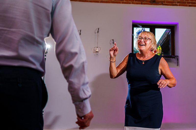 An elderly woman with blonde hair and glasses joyfully dances in a well-lit room with purple accents. A man in a white shirt and dark pants faces her, slightly blurred in the foreground. Both appear to be enjoying the moment. Image by Documentary Wedding Photographer Lewis Bishop Photography.