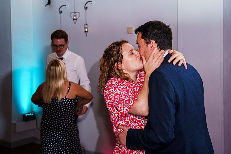 A couple shares a kiss while dancing at an indoor event. The woman is wearing a red-patterned dress, and the man is in a dark suit. In the background, another couple is dancing, with blue accent lighting on the wall and decorative hangings above. Image by Documentary Wedding Photographer Lewis Bishop Photography.