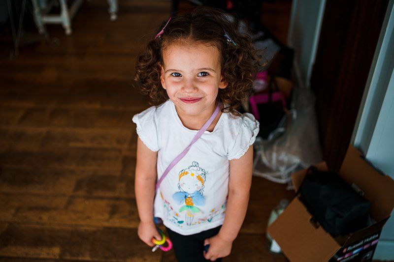 A young girl with curly hair and a bright smile looks up at the camera. She is wearing a white shirt with a colorful illustration on it and is holding a toy. The background includes a wooden floor and some scattered items, including a cardboard box. Image by Documentary Wedding Photographer Lewis Bishop Photography.