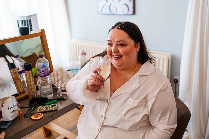 A woman with dark hair smiles while holding a glass of champagne in a light-filled room. She is sitting in front of a vanity table with various personal items, including a water bottle, hair products, and a purse. She wears a white button-down shirt. Image by Documentary Wedding Photographer Lewis Bishop Photography.