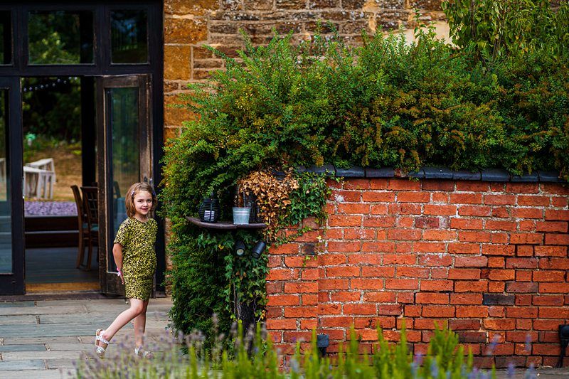 A young girl in a patterned dress stands near an outdoor brick structure with greenery on top, smiling and looking to the side. Behind her is a building with a large glass door. The scene is vibrant and colorful, with a peaceful and natural atmosphere. Image by Documentary Wedding Photographer Lewis Bishop Photography.