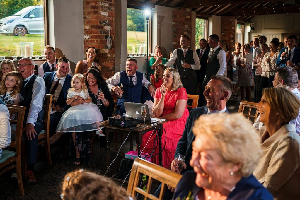 A lively gathering in a rustic venue with large windows and exposed brick walls. Adults and children, dressed in formal attire, are seated, smiling, and laughing. Lights illuminate the room, enhancing the festive and joyful atmosphere. Image by Documentary Wedding Photographer Lewis Bishop Photography.