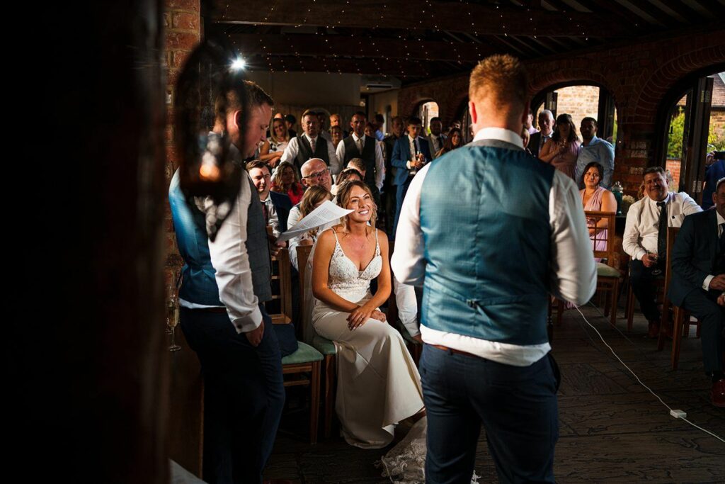 A bride in a white gown sits smiling, facing a man in a blue vest speaking in front of an audience. Fairy lights decorate the ceiling. The onlooking crowd, dressed in formal attire, appears engaged and festive in the rustic, warmly lit venue. Image by Documentary Wedding Photographer Lewis Bishop Photography.