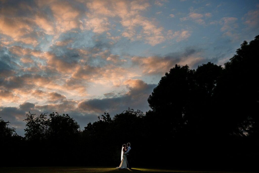 A bride and groom stand closely together in an open field, silhouetted against a vibrant sunset sky filled with dramatic clouds. Tall trees frame the scene, creating a picturesque and romantic atmosphere. Image by Documentary Wedding Photographer Lewis Bishop Photography.