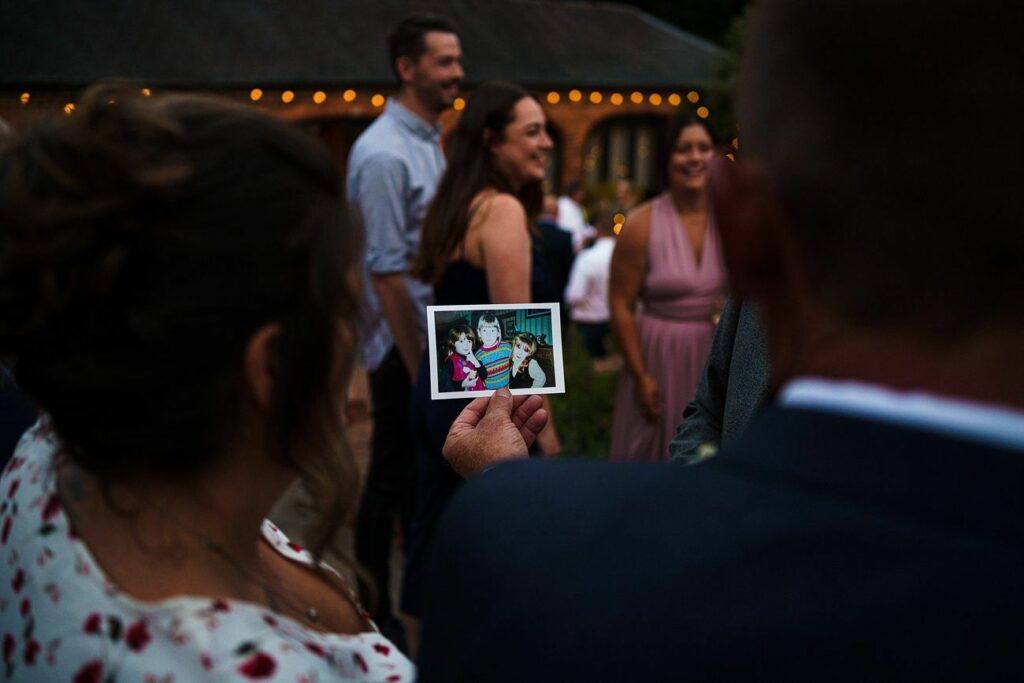 A group of people are gathered outdoors in the evening, dressed in semi-formal attire. One person is holding a photo of two children. String lights are visible in the background, creating a warm, festive atmosphere. The focus is on the hand holding the photo. Image by Documentary Wedding Photographer Lewis Bishop Photography.