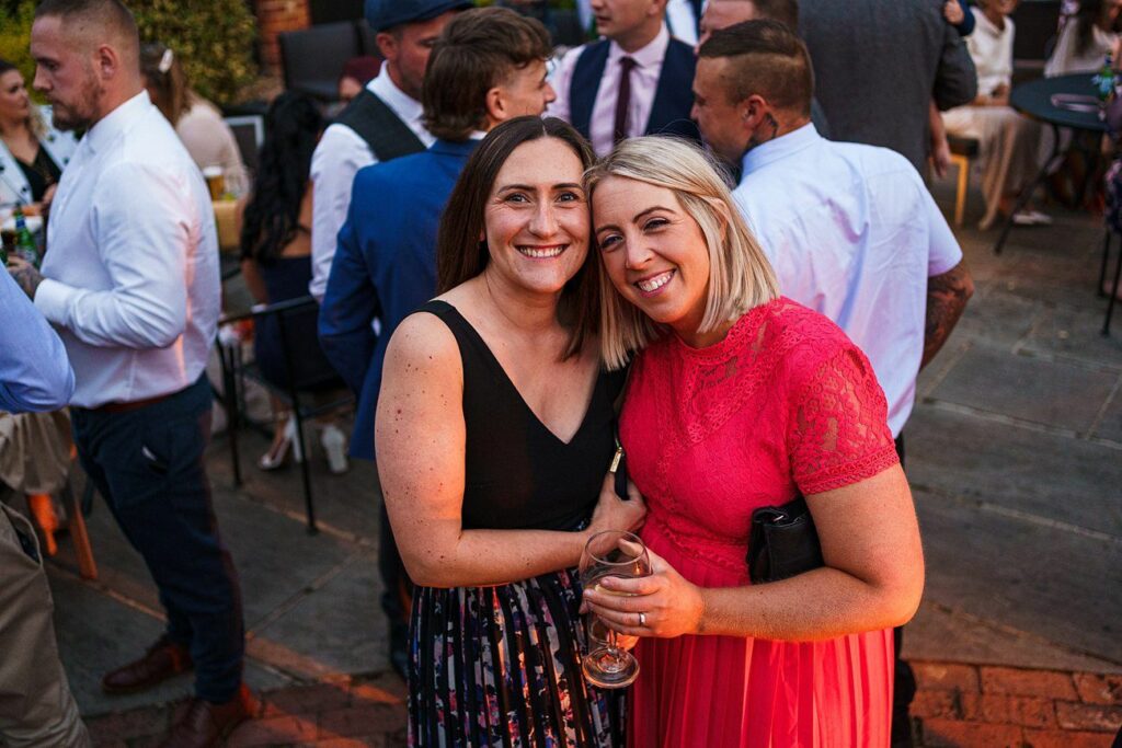 Two women are smiling at the camera in an outdoor social gathering. One wears a black top and patterned skirt, and the other wears a red lace dress while holding a drink. Other people in the background are mingling. The event seems to be warm and lively. Image by Documentary Wedding Photographer Lewis Bishop Photography.