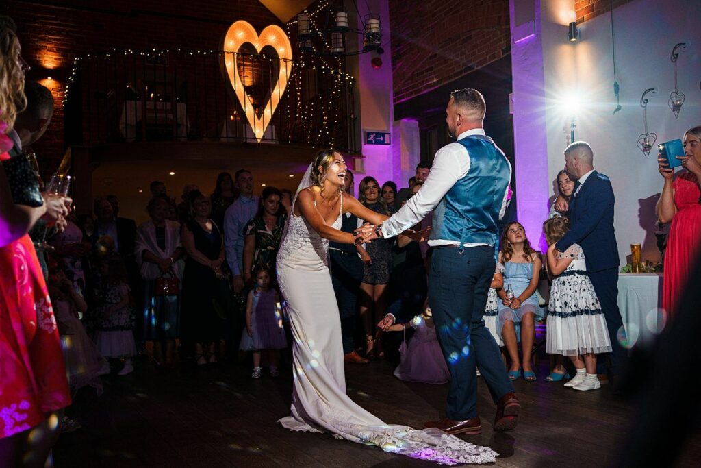 A bride in a white dress and a groom in a blue vest enjoy their first dance at their wedding. They are surrounded by guests watching them, and there is a large heart-shaped light fixture in the background. The room has ambient purple lighting. Image by Documentary Wedding Photographer Lewis Bishop Photography.