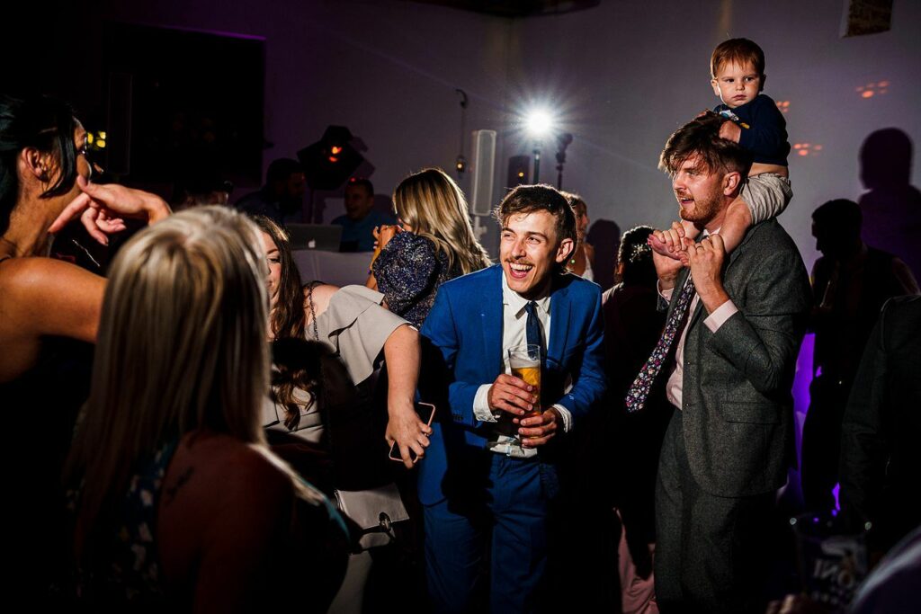 A group of people are dancing and laughing at a party. One man in a blue suit is holding a drink and smiling broadly. Another man holds a toddler on his shoulders. The background is dark with colorful lighting that creates a lively atmosphere. Image by Documentary Wedding Photographer Lewis Bishop Photography.