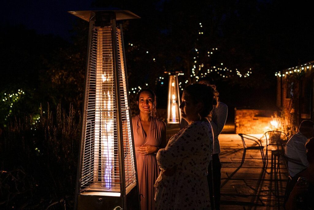 Several people stand outdoors at night, gathered around a tall outdoor heater with visible flames. String lights illuminate the background, giving the setting a warm and festive ambiance. One person in a light pink dress is smiling while others wear light jackets. Image by Documentary Wedding Photographer Lewis Bishop Photography.