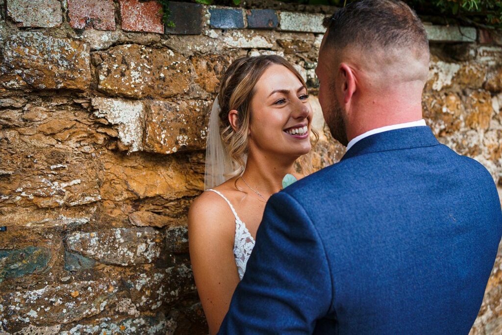 A newlywed couple stands close to each other in front of a rustic stone wall. The bride, wearing a white lace dress and veil, smiles warmly at the groom, who is dressed in a blue suit, as they share an intimate moment. Image by Documentary Wedding Photographer Lewis Bishop Photography.