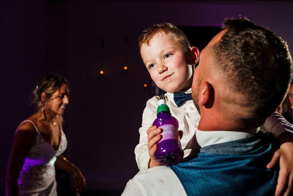 A young boy in a bow tie is being held by an adult man in a blue vest. The boy is holding a small purple bottle and looking to the side. In the background, a woman in a white dress, presumably a bride, is smiling and watching them. The setting appears to be a wedding reception. Image by Documentary Wedding Photographer Lewis Bishop Photography.
