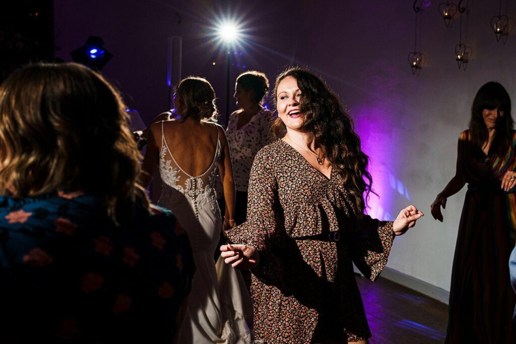 A group of people, dressed in casual and formal attire, dance in a dimly lit room with colorful lighting. A woman with long curly hair and wearing a floral dress is in the foreground, smiling and dancing joyfully. A bright light shines from the background. Image by Documentary Wedding Photographer Lewis Bishop Photography.
