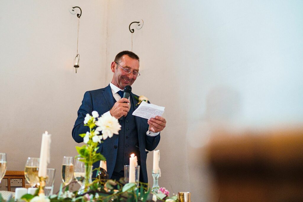 A man in a blue suit and glasses holds a microphone and a piece of paper, smiling as he delivers a speech. In the foreground are lit candles, flowers, and champagne glasses, suggesting a celebratory event. The environment appears bright with a light-colored wall behind him. Image by Documentary Wedding Photographer Lewis Bishop Photography.