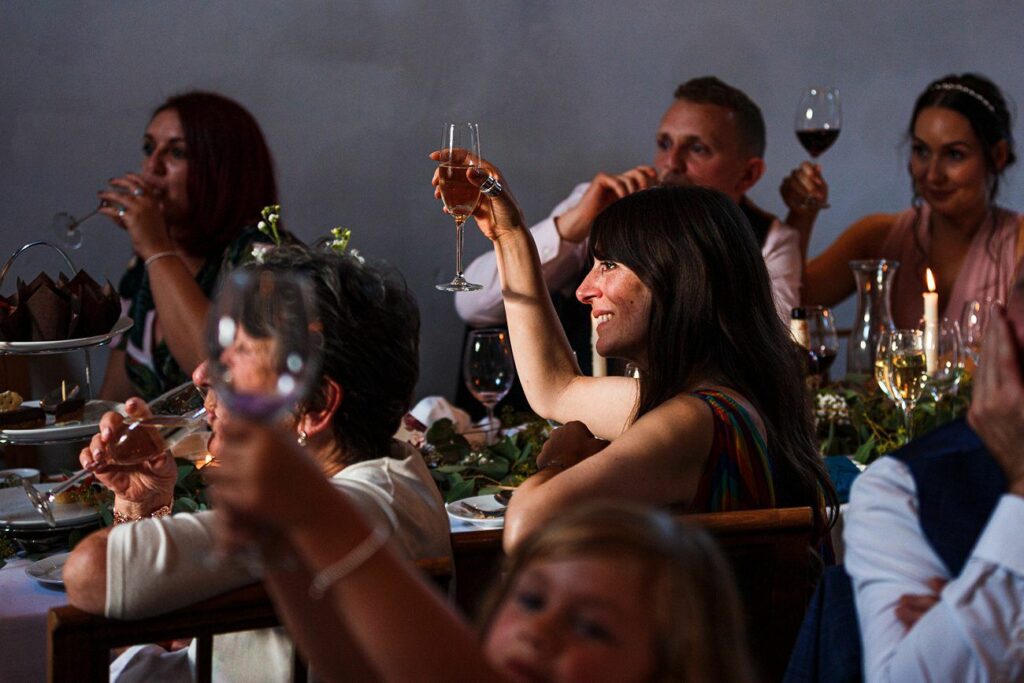 A group of people raising their glasses in a toast at a dinner party. The table is set with food, drinks, and decorative greenery. A woman in the center, smiling, holds her glass up, reflecting warm light on her face. The atmosphere is celebratory and joyful. Image by Documentary Wedding Photographer Lewis Bishop Photography.