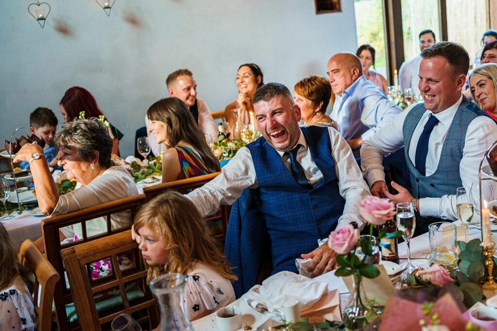 A group of people, dressed in formal attire, are seated at tables in a well-lit room. They are laughing, talking, and enjoying themselves. The table in the foreground has flowers, drink glasses, and a young girl with blonde hair at the end. 

. Image by Documentary Wedding Photographer Lewis Bishop Photography.