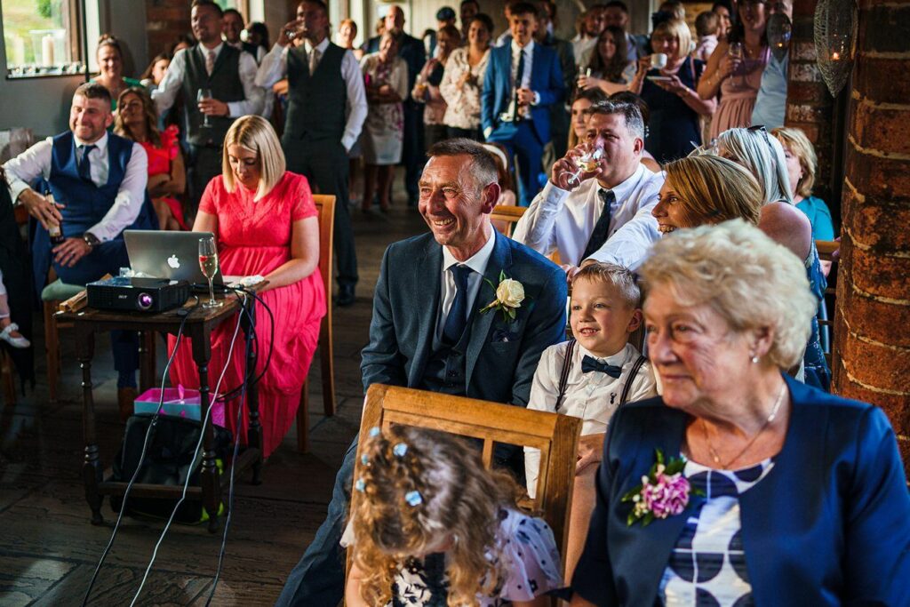 A diverse group of people is gathered at an indoor event, seated and smiling. The audience includes men, women, and children, dressed semi-formally. A projector is set up on a table. The atmosphere appears joyful and relaxed, with many people smiling or laughing. Image by Documentary Wedding Photographer Lewis Bishop Photography.