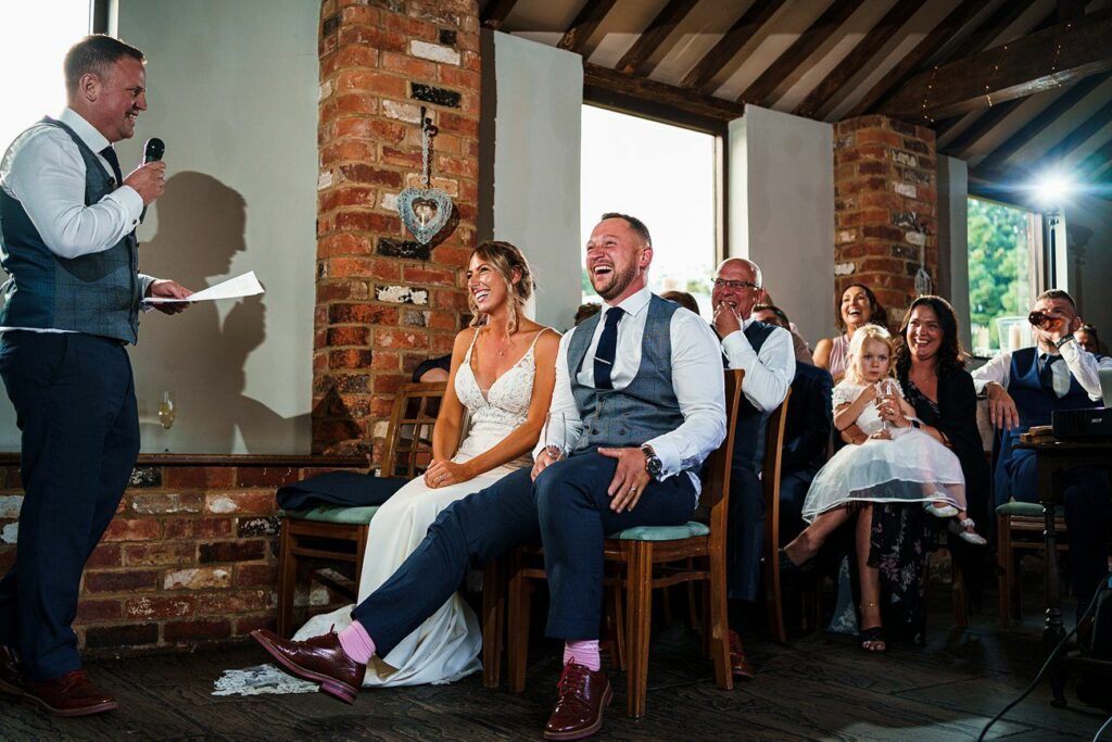 A bride and groom sit smiling and laughing as a man in a vest and tie gives a speech. The venue has brick walls and large windows. Guests, including children, are seated around them, equally engaged and joyful. The scene is warm and lively. Image by Documentary Wedding Photographer Lewis Bishop Photography.