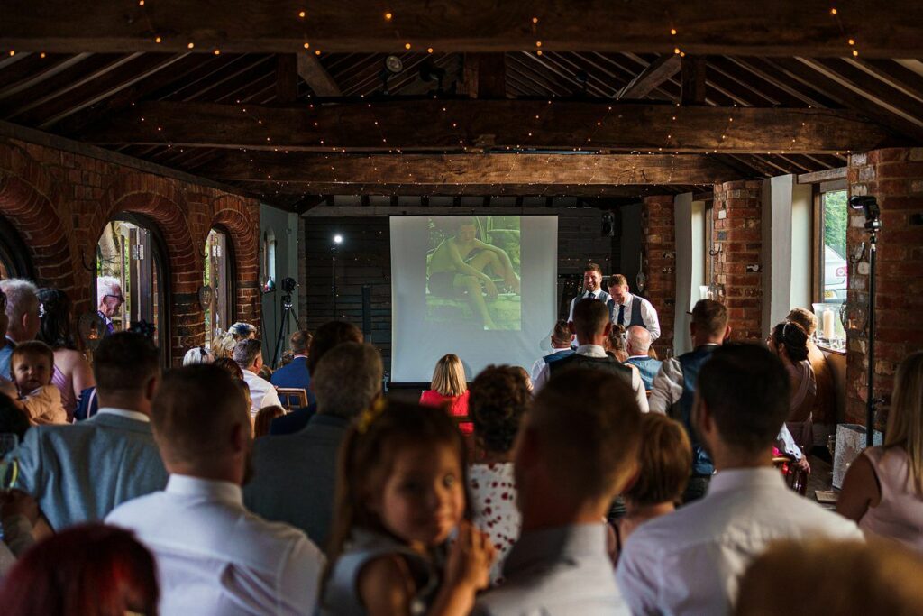 A group of people gathered in a rustic hall with brick walls and wooden beams, watching a presentation projected on a screen. The image on the screen shows a couple embraced in a romantic setting. Twinkling lights are strung above, creating a warm atmosphere. Image by Documentary Wedding Photographer Lewis Bishop Photography.
