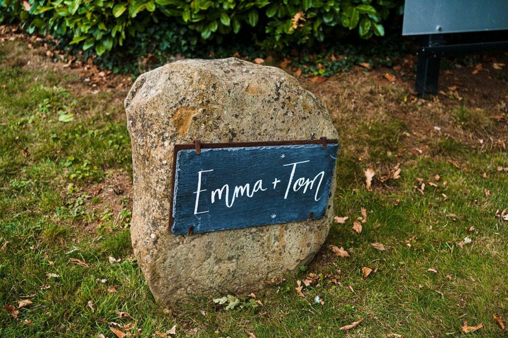 A large rock placed in a grassy area has a dark rectangular plaque attached to it. The plaque is inscribed with the names "Emma + Tom" in white, cursive writing. Green foliage is in the background. Image by Documentary Wedding Photographer Lewis Bishop Photography.
