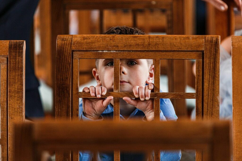 A young child with short brown hair peers through the wooden lattice backrest of a chair. The child is wearing a light blue shirt and appears to be in an indoor setting, surrounded by similar wooden chairs. Their hands are gripping the chair's backrest. Image by Documentary Wedding Photographer Lewis Bishop Photography.