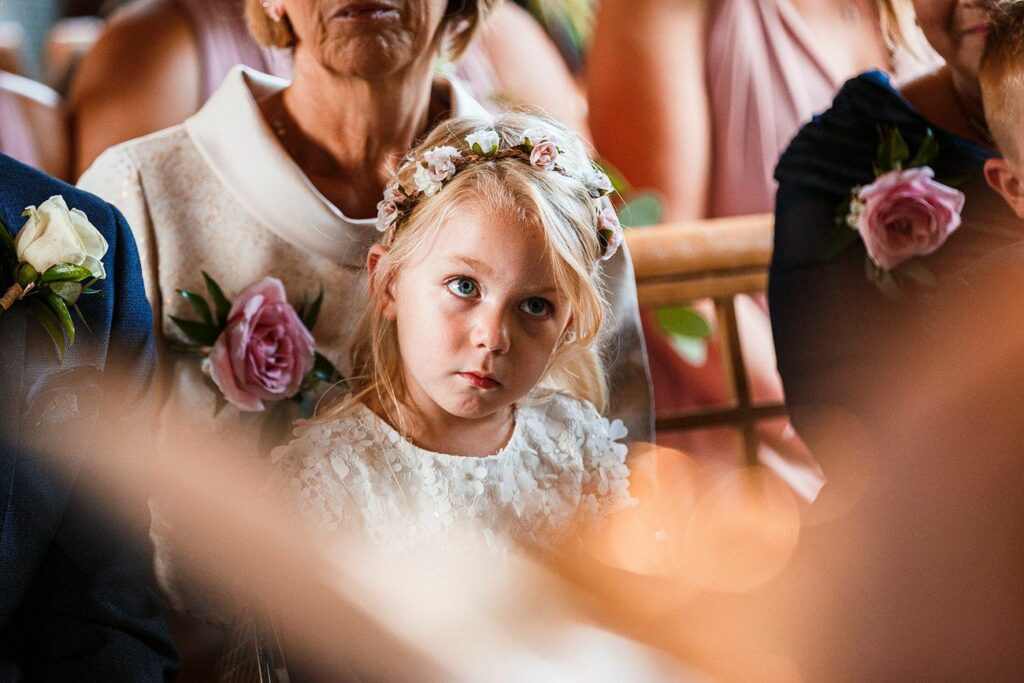 A young girl with blonde hair, wearing a white dress and a floral headband, sits among adults at what appears to be a formal gathering or ceremony. She looks slightly up with a serious expression. The adults around her are dressed in formal attire with flower boutonnieres. Image by Documentary Wedding Photographer Lewis Bishop Photography.