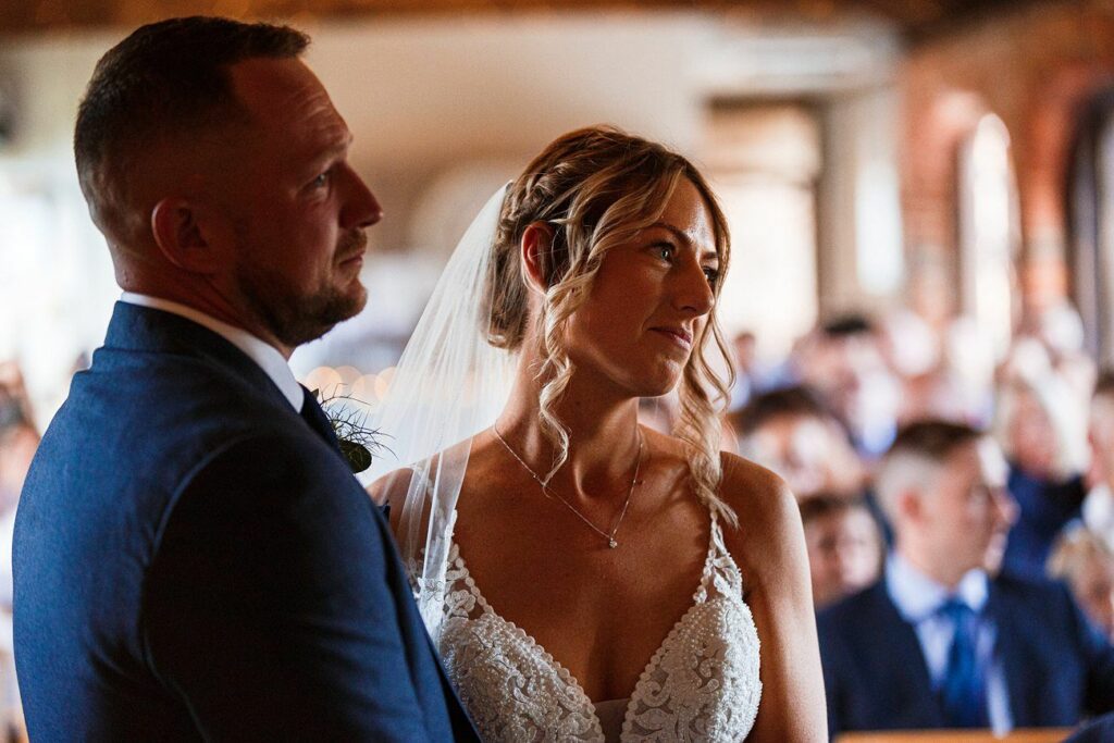 A couple, with the groom in a blue suit and bride in a white lace dress with a veil, stand close to each other in a warmly lit room with guests in the background. The bride is looking to the side with a gentle smile. Image by Documentary Wedding Photographer Lewis Bishop Photography.