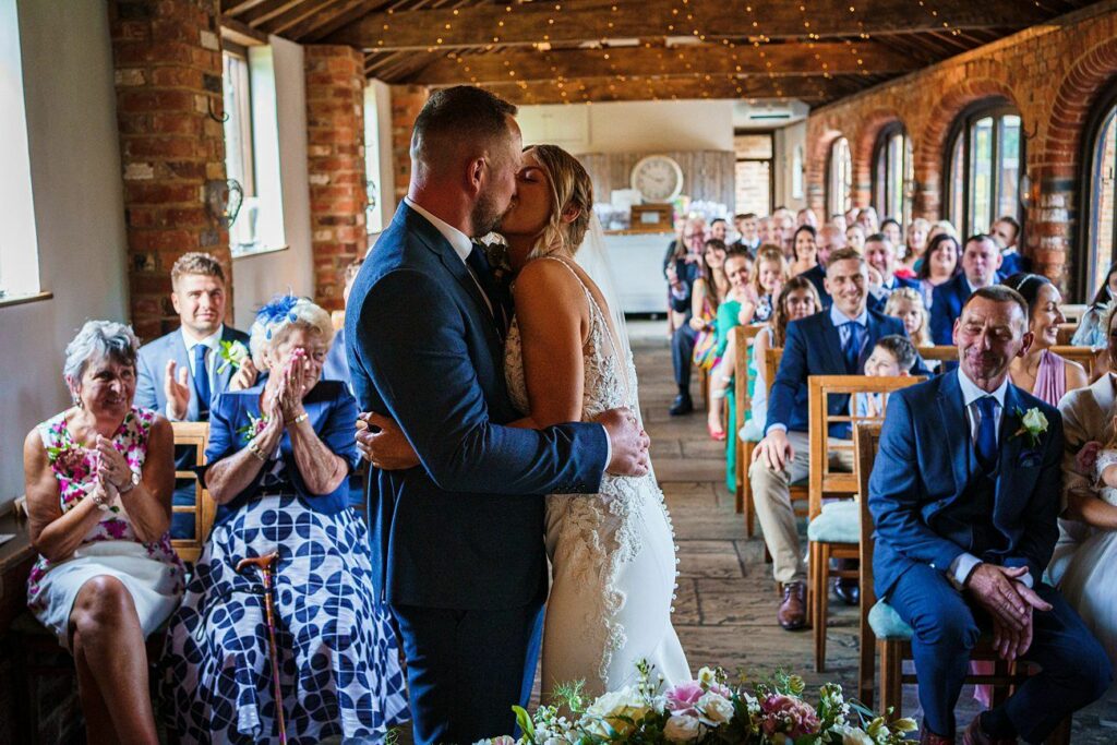 A bride and groom kiss at the altar in a warmly lit, rustic wedding venue with exposed brick walls and wooden beams. Guests, seated in rows, watch and clap. Some have broad smiles, showing joy and celebration. Flowers adorn the altar in the foreground. Image by Documentary Wedding Photographer Lewis Bishop Photography.