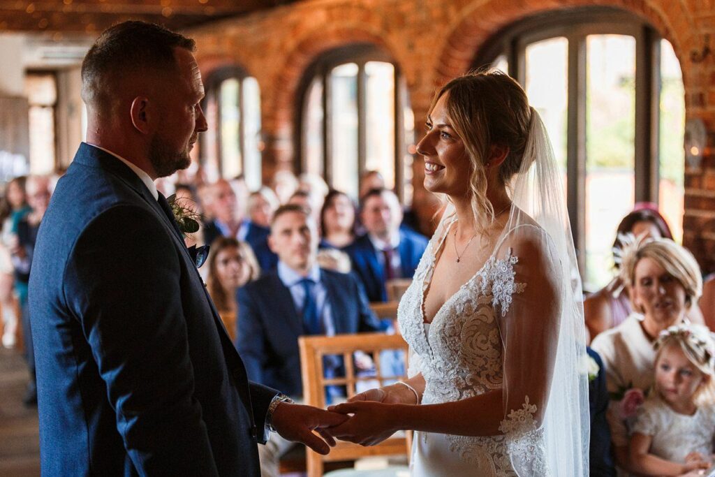 A bride and groom stand facing each other, holding hands during their wedding ceremony inside a warmly lit room with brick walls and arched windows. The bride is in a lace wedding gown and veil, and the groom is in a dark suit. Guests seated in the background watch them. Image by Documentary Wedding Photographer Lewis Bishop Photography.