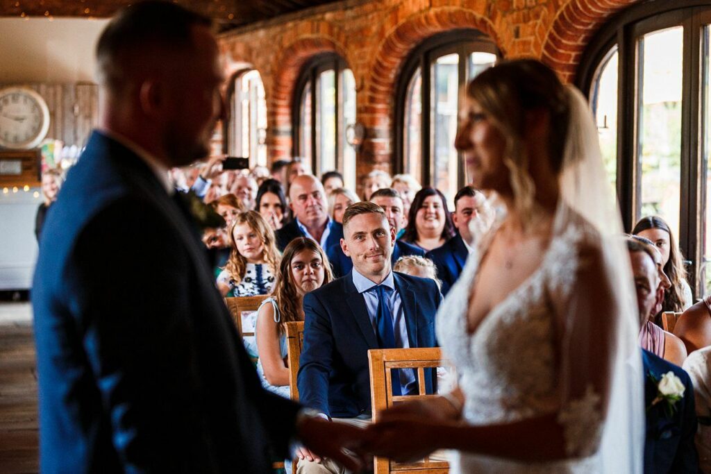 A couple stands face-to-face holding hands during their wedding ceremony in a warmly lit, rustic venue with exposed brick walls and large windows. Guests sit in rows of chairs, attentively watching the ceremony, some smiling. Image by Documentary Wedding Photographer Lewis Bishop Photography.