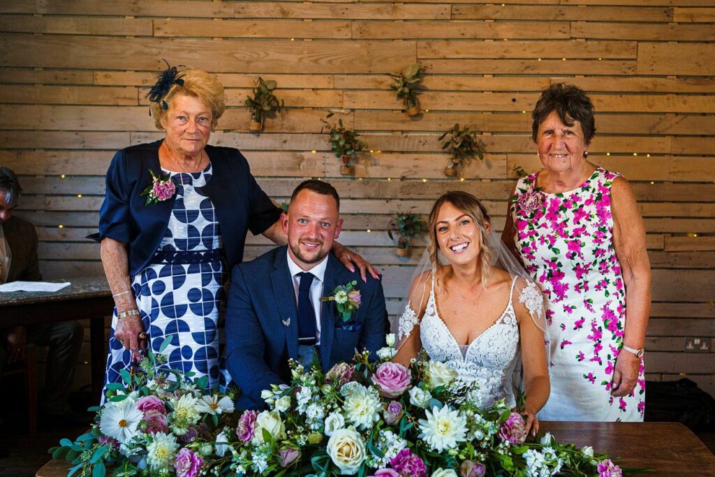 A bride and groom pose for a photo with two older women at their wedding. The group stands in front of a wooden backdrop adorned with hanging greenery. A floral arrangement is placed in front of the couple, who are seated and smiling. Image by Documentary Wedding Photographer Lewis Bishop Photography.