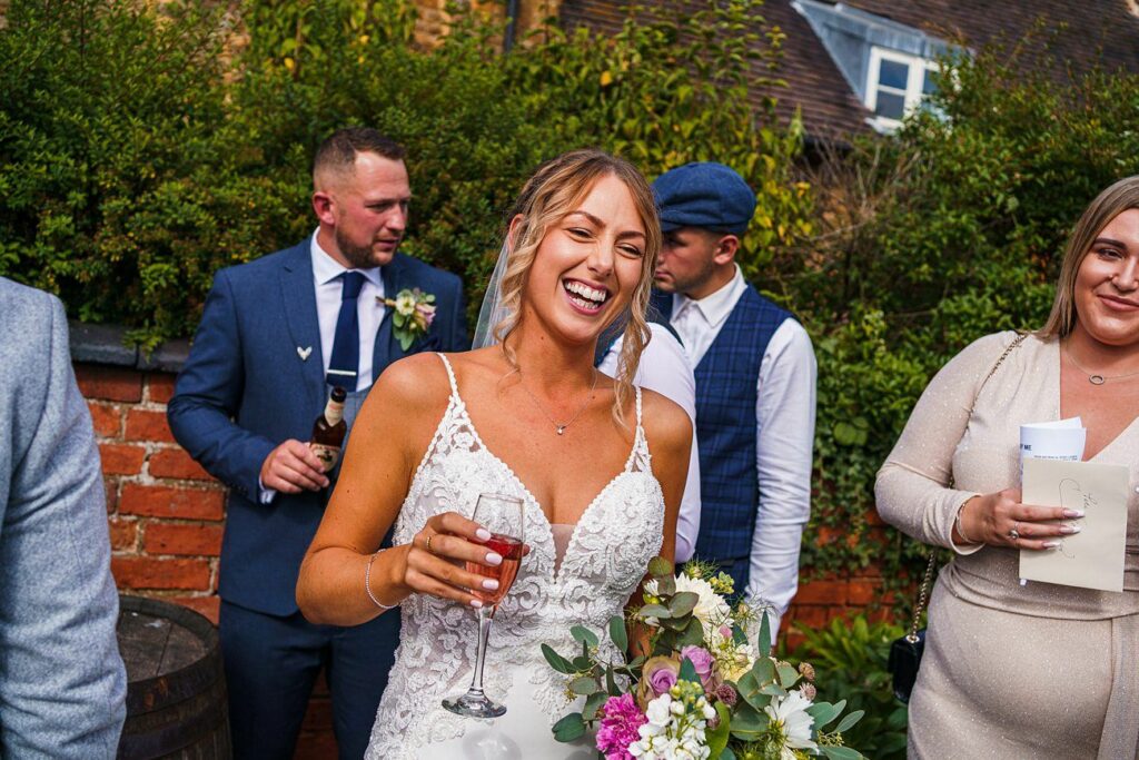 A smiling bride in a white lace dress holds a glass of rosé wine and a bouquet of flowers. She is outside, with three casually dressed guests in the background. The group is standing near a brick wall, with greenery and a house visible behind them. Image by Documentary Wedding Photographer Lewis Bishop Photography.