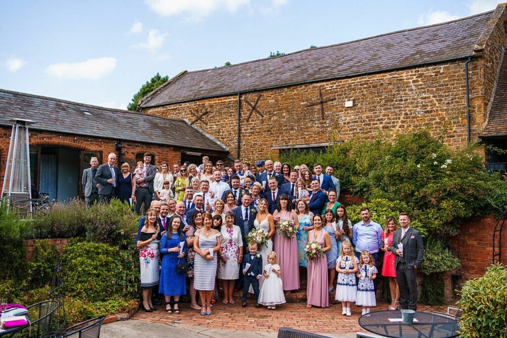 A large group of people dressed in formal attire gather outdoors for a wedding photo. The group includes the bride, groom, bridesmaids, groomsmen, and many guests, standing in front of a rustic brick building with greenery around. Some children are in the front. Image by Documentary Wedding Photographer Lewis Bishop Photography.