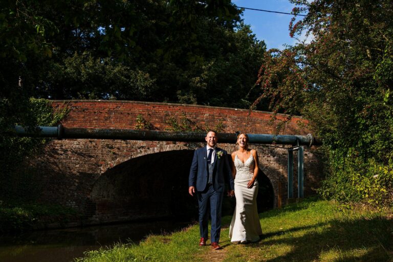 A couple dressed in wedding attire stands outdoors in front of a brick bridge. The groom is in a navy suit with a boutonnière, and the bride is in a white dress. They are holding hands and smiling, surrounded by lush greenery and grass. Image by Documentary Wedding Photographer Lewis Bishop Photography.