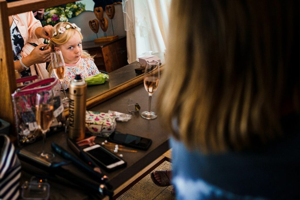 A young girl with a flower crown looks at her reflection in a mirror while having her hair styled by an adult. Various beauty supplies, such as a curling iron, hairspray, and makeup, are scattered on the vanity. A glass of rosé wine is also on the table. Image by Documentary Wedding Photographer Lewis Bishop Photography.