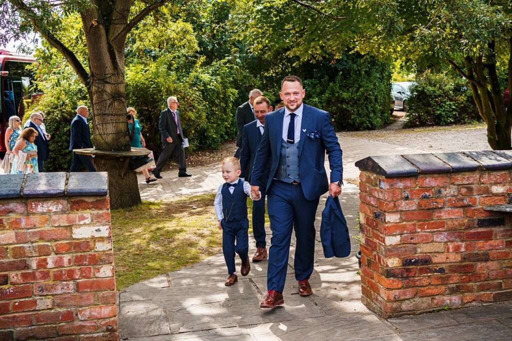 A man in a blue suit holding a small child's hand walks along a stone path. Both are dressed formally and smile as they approach a brick wall entrance. Several other individuals, also dressed formally, can be seen in the background near trees and greenery. Image by Documentary Wedding Photographer Lewis Bishop Photography.
