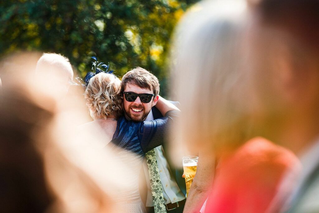 A man in sunglasses with a beard hugs a woman in a blue dress at an outdoor gathering. The man is smiling and holding a drink. The background features trees and sunlight, and out-of-focus people surround them, creating a warm, festive atmosphere. Image by Documentary Wedding Photographer Lewis Bishop Photography.