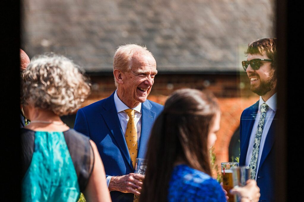 A group of people dressed in formal attire is gathered outdoors, engaging in conversation. An elderly man in a blue suit and yellow tie is smiling warmly, holding a drink, while a younger man with sunglasses stands next to him, also smiling. Image by Documentary Wedding Photographer Lewis Bishop Photography.